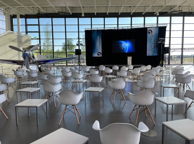 Heller Veranstaltungsraum mit weißen Designerstühlen, ausgerichtet auf eine Bühne mit Leinwand und Rednerpult, große Glasfassade mit Blick auf Flugzeuge und Außengelände.