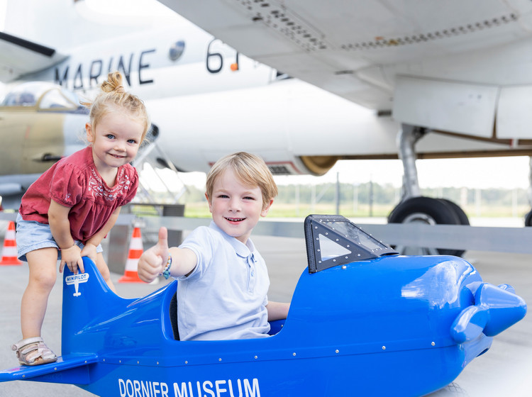 Zwei kleine Kinder sitzen auf einem Spielzeugflugzeug in einem Hangar, im Hintergrund steht ein großes Flugzeug.