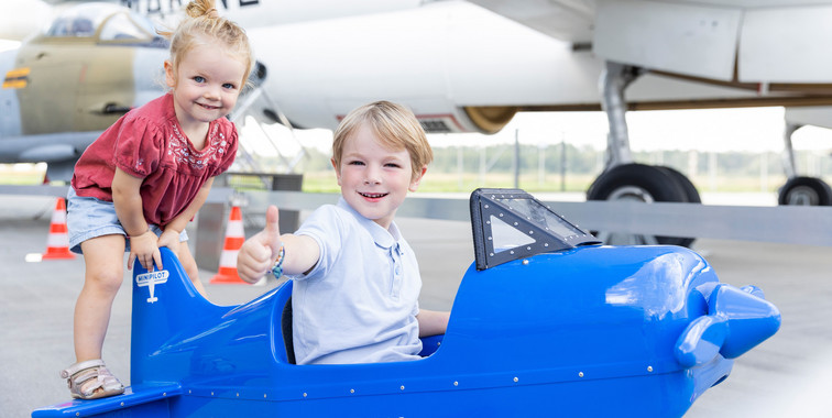 Zwei kleine Kinder sitzen auf einem Spielzeugflugzeug in einem Hangar, im Hintergrund steht ein großes Flugzeug.