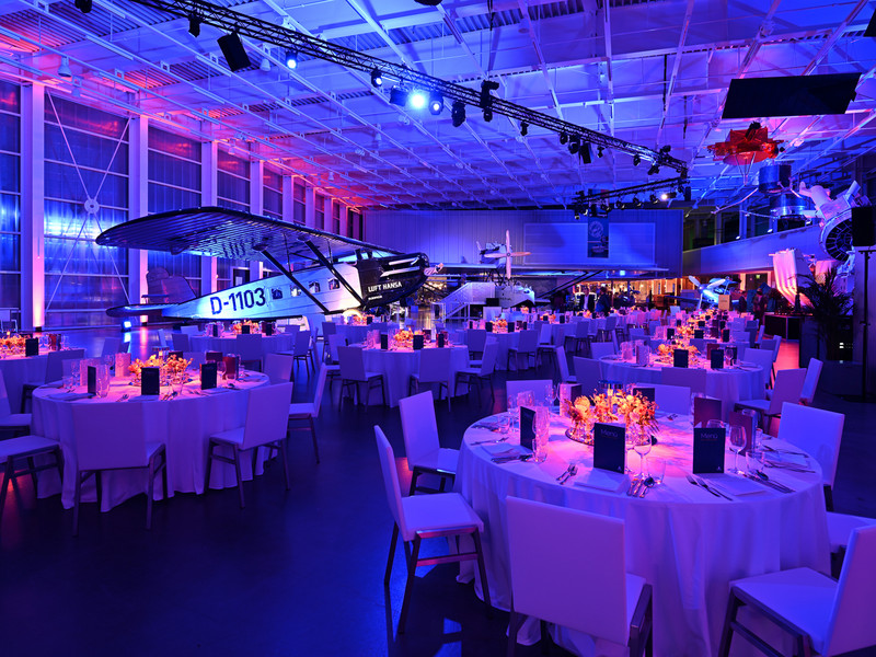 Eventlocation mit runden, weiß gedeckten Tischen in stimmungsvoller Blau- und Violettbeleuchtung, Flugzeuge als Dekorationselemente unter der Decke.
