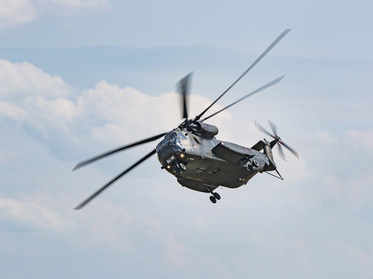 Großer Militärhubschrauber mit laufenden Rotoren im Flug vor leicht bewölktem Himmel.