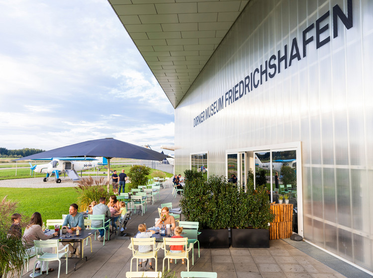 Mehrere Personen sitzen an den Tischen im Außenbereich des Dornier-Restaurants mit dem Rollfeld im Hintergrund.