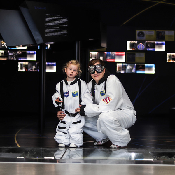 Zwei Kinder in weißen Astronautenanzügen stehen und knien vor einer dunklen Raumfahrtausstellung mit beleuchteten Bildtafeln.