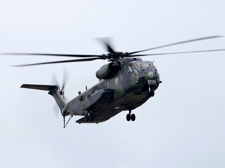 Grauer Militärhubschrauber mit ausgefahrenem Fahrwerk und rotierenden Rotorblättern im Flug vor hellem Himmel.