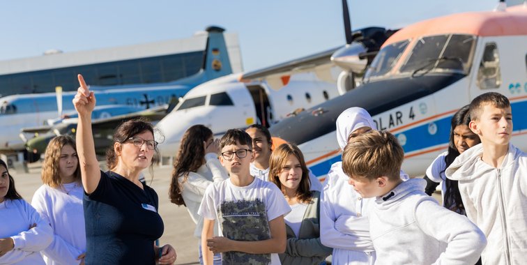 Im Vordergrund stehen Schüler während im Hintergrund mehrere Flugzeuge auf dem Rollfeld stehen.