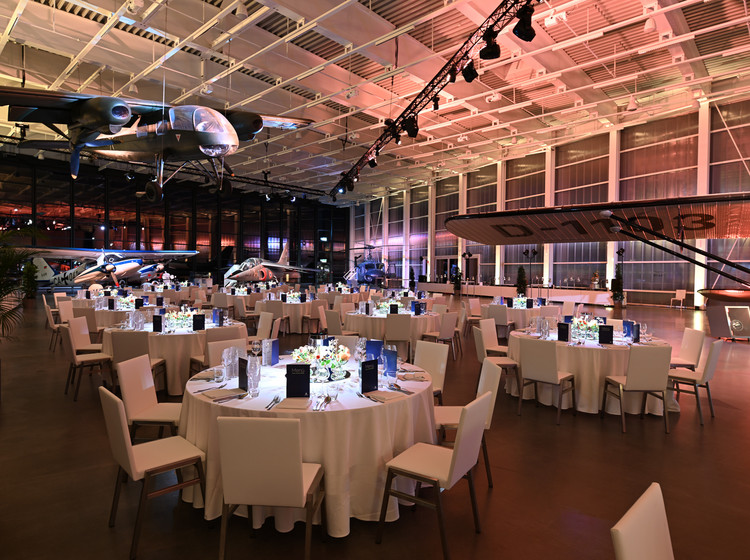 Stimmungsvoll beleuchtete Eventlocation mit runden, festlich gedeckten Tischen, weißer Bestuhlung und großer Glasfassade, im Hintergrund Flugzeuge und Beleuchtung in warmen Farben.