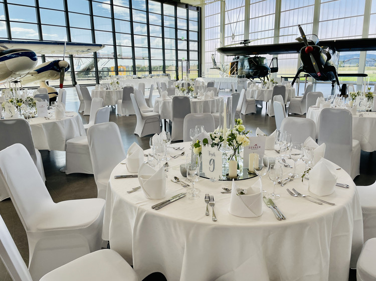 Rund eingedeckter Tisch mit weißen Hussenstühlen, Besteck, Gläsern und Blumendeko in lichtdurchfluteter Eventlocation mit Flugzeugen vor großer Glasfront.