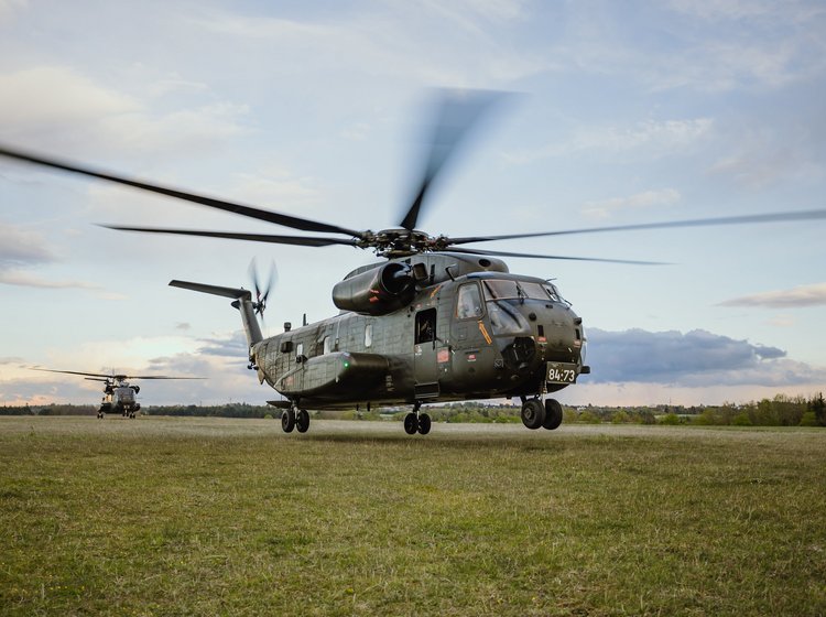 Grauer Militärhubschrauber mit laufendem Rotor auf einer Wiese unter bewölktem Himmel.