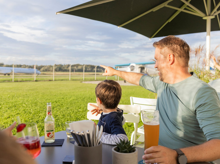 Ein Vater sitzt mit seinem Sohn im Außenbereich des Dornier-Restaurants am Tisch und zeigt auf das Rollfeld im Hintergrund.