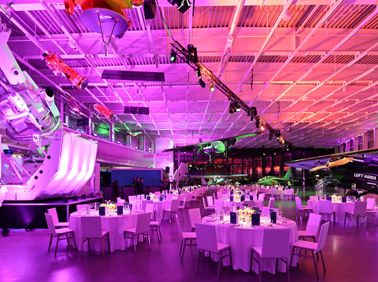 Große Eventhalle mit runden Tischen und weißer Bestuhlung in pink-violetter Lichtstimmung, dekorativer Bühne und aufgehängtem Flugzeug über dem Veranstaltungsbereich.