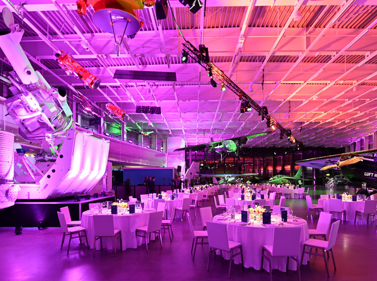 Große Eventhalle mit runden Tischen und weißer Bestuhlung in pink-violetter Lichtstimmung, dekorativer Bühne und aufgehängtem Flugzeug über dem Veranstaltungsbereich.